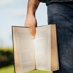 Crop unrecognizable male student in casual outfit standing with book in hand on green spring meadow with sunshine lighting up pages of novel